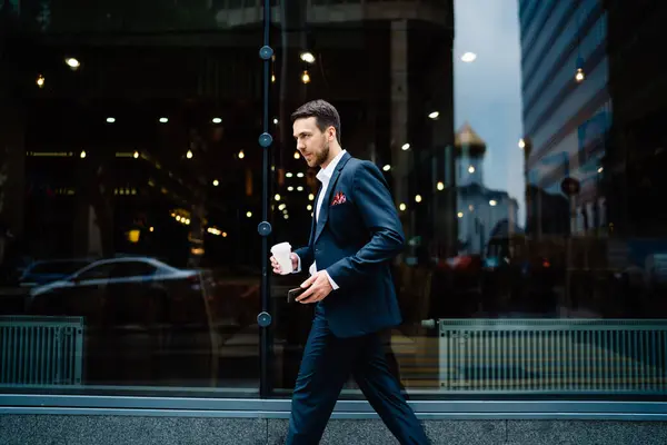 Side view of content manager in dark elegant suit with pocket square in pocket and mobile phone walking along black windows reflecting city lights in downtown
