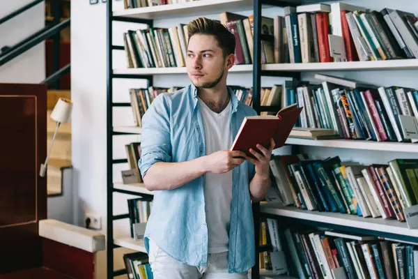 Kitap raflarına yaslanan zeki bir öğrenci ve açık bir kitapla sayfayı çevirirken başka tarafa bakıyor ve hafif kütüphanede seçiyor. 