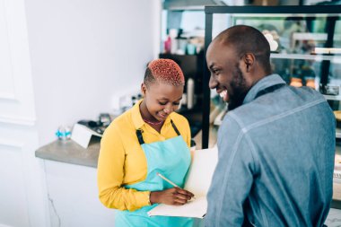 Afro-Amerikalı genç, önlüklü, neşeli meslektaşları şehirdeki kafeteryada birbirleriyle sohbet ederken deftere not alıyorlar.