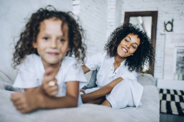 Beyaz elbiseli oyuncu kıvırcık saçlı anne aynı kıza dokunuyor stüdyoda yatakta oturuyor ve arka planda büyük bir ayna var.