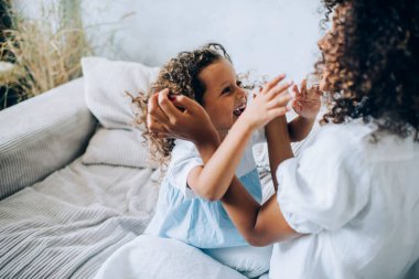 Beyaz elbiseli, etnik kıvırcık saçlı kadını kandırmak için yukarıdan. Küçük sevimli bir kızla yatakta oturmuş stüdyo dairesinde birbirlerinin saçlarıyla oynuyorlar. 