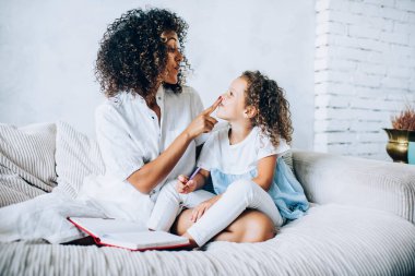 Beyaz elbiseli mutlu etnik anne yatakta kıvırcık saçlı kızı burnuna dokunarak evde kitap okuyor.