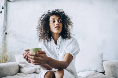 Düşünceli, barışçıl, çekici, Afro saçlı, ev istirahatı sırasında her iki elinde de bir kupa sıcak içecekle etnik kadın.