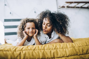 Etnik kıvırcık saçlı, sıradan giyinmiş, hikaye anlatan bir anne. Bebek yatakta uzanıp kameraya bakıyor.