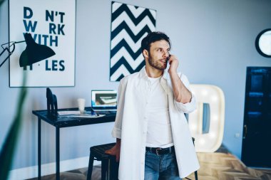 Beyaz tişörtlü, beyaz tişörtlü neşeli erkek yaratıcı iş yerindeki akıllı telefondan çağrı yapıyor. Duvarında siyah beyaz resimler var.