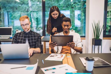 Gülümseyen siyah öğrenci, gözlüklü kızıl saçlı erkek arkadaşı yanında otururken ve dizüstü bilgisayar kullanırken kadın grup arkadaşına tablet ekranı gösteriyor.