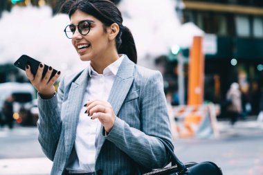 Yuvarlak gözlüklü, şık, klasik takım elbiseli, neşeli ve iyimser bir bayan çalışan sokakta tatlı bir gülümsemeyle yürüyor ve telefonda konuşuyor.