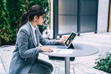 Kendine güvenen, kendine güvenen, zarif giyinen, gözlüklü, at kuyruklu, kafeteryanın dışında tablet üzerinde sörf yapan güzel bir kadının yan görüntüsü.