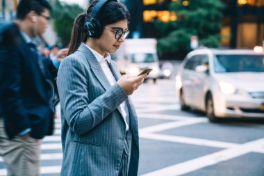 Modern kadın çalışan, gözlüklü ve takım elbiseli. Kablosuz kulaklıklarla işlek şehir caddesinde dururken akıllı telefon kullanıyor.