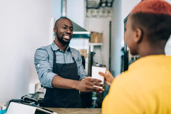 Young cheerful African American employee in black apron standing at counter in small shop and serving coffee takeaway for female customer