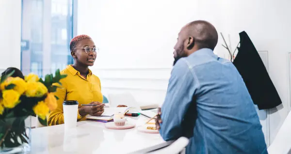 African American gorgeous female in yellow shirt and glasses with pen in hand writing memo in notepad working on project with black male eating dessert drinking coffee in pastry shop