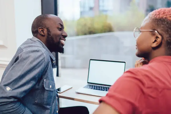 Positive black guy in casual clothes creating startup using laptop discussing strategy with African American crop lady in office looking at each other