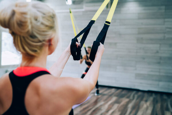 Back view of slim woman with blonde hair in sport clothes standing and exercising with rubber expander in fitness club