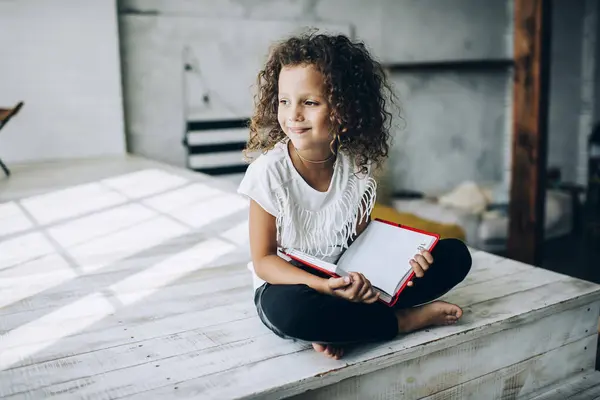 Stock image Thinking smiling adorable curly haired kid in casual apparel reading book sitting with crossed legs on wooden podium in light studio in sunny day