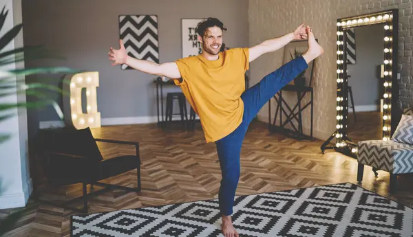 Optimistic guy in casual wear with raised leg standing on one leg and stretching doing yoga exercise practicing meditation and healthy lifestyle in modern flat