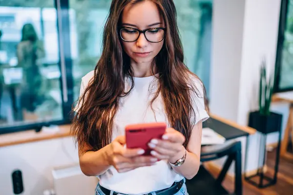 Young female in glasses and casual clothes browsing modern smartphone while standing against table and window during work in creative workspace
