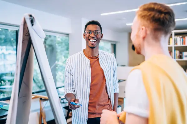 Cheerful black man pointing at flip chart and laughing at joke while working with blurred young man in modern creative workspace
