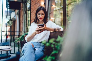 Beyaz gömlekli modern bir bayan cep telefonuyla mesajlaşan yırtık kot pantolonlu açık hava terasında masada oturuyor.