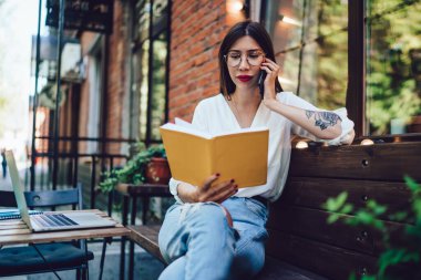 Ciddi şekilde odaklanmış, beyaz gömlekli, cep telefonuyla konuşan, yazın açık terasta oturan not defterine bakan,