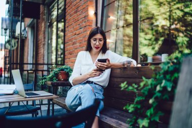 Mutlu, modern, beyaz gömlekli, yırtık kot pantolonlu genç bayan yazın kafede cep telefonuyla mesajlaşıyor.