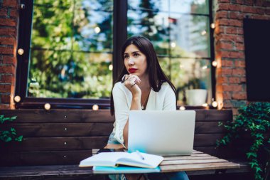 Düşünceli İspanyol milenyumu kafeterya masasında oturuyor ve sosyal medyada web yayını fikrini düşünürken başka tarafa bakıyor, modern teknolojiyi kullanan kadın nesli Y