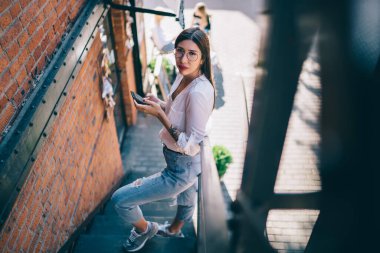 Gözlüklü, beyaz bluzlu, spor ayakkabılı, yangın merdiveninde dikilip kameraya bakarken cep telefonunu karıştıran Asyalı bir kadın.