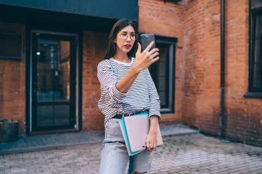 Konsantre olmuş genç esmer, cep telefonuyla kendi portresini çekerken elinde dizüstü bilgisayar ve tuğlalı şehir evinin yanında duruyor.