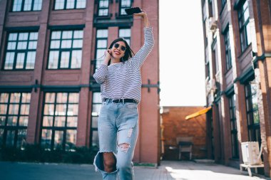 Neşeli ruh hali içinde neşeli bir hipster kızı açık havada müzik podcasından dans ederken heyecanlı ve mutlu. Elektronik kulaklıklar ve nesiller boyu akıllı telefon uygulamaları ile dinleyin.