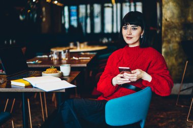 Elinde cep telefonu olan neşeli ve sakin bir kadın iş sırasında dizüstü bilgisayarını düşünüyor ve kafede gözlerini kaçırarak not defterini açıyor.