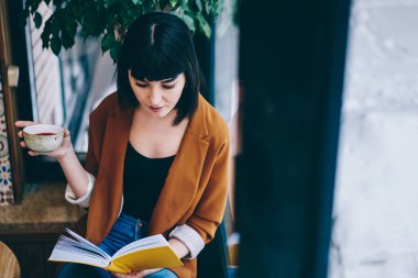 Yukarıdan bir fincan kırmızı çayla şık bir kadın pencerenin yanında oturmuş restoranda dinlenirken ilginç bir kitap okuyor.