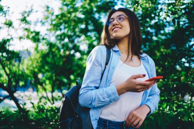 Güneşli bir günde, güneşli bir günde eğlenen neşeli, gözlüklü genç hippi kızla parkta cep telefonu ve 4G bağlantısı kullanarak gezen güzel bir kadın.