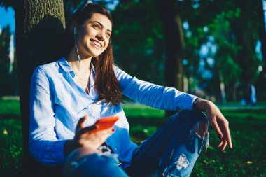 Parkta ağaçların yanında oturan güzel hippi kız, cep telefonuyla kulaklıkla müzik dinliyor, milenyum kadınlarına gülümsüyor güneşli bir günde cep telefonu ve aksesuarla dinlenmekten ve internetten radyo dinlemekten keyif alıyor.