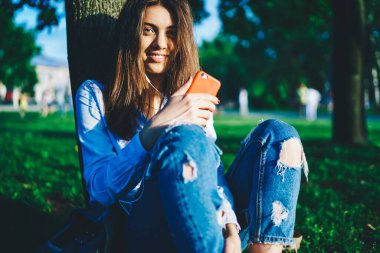 Beyaz saçlı çekici bir kadın kameraya bakıyor, akıllı telefondaki uygulamadan sesli kitap dinliyor, cep telefonu ve kulaklık kullanarak güneşli bir günde şarkı çalmaktan zevk alan bin yıllık kadın portresi.