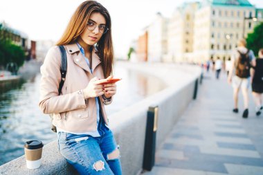 Nehir köprüsünde cep telefonu kullanarak mesajlaşan ve açık havada posta gönderen modaya uygun giyinmiş yeniyetme kız, genç bayan seyahat blogcusu şehir ortamlarında akıllı telefon ile medyayı paylaşıyor