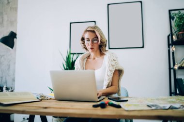 Güzel, sarışın, gözlüklü, ağzı açık, iş yerinde fotoğraf makineleri ve çizimlerle dolu bir laptop kullanan serbest çalışan bir kadın.