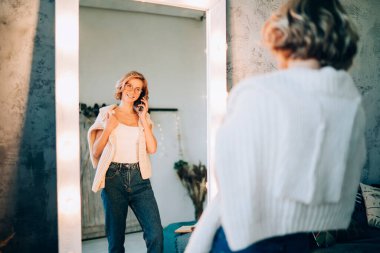 Optik gözlüklü mutlu bir kadın soyunma odasındaki lamba aynasının yanında poz veriyor ve moda tasarımcısıyla cep telefonu konuşması yapıyor. Moda kıyafetlerini tartışıyor, telefonla gülümseyen bir kadın arıyor.