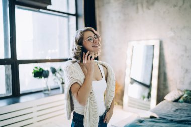 Beyaz süveter ve gözlüklü neşeli sarışın kadın. Parlak ışıklı yatak odasında el ele tutuşup telefonla konuşuyor.