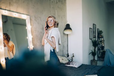 Gözlüklü, parlak ve neşeli genç bayan. Aydınlanmış aynanın önünde duruyor. Geniş yataklı ve telefonla konuşuyor.