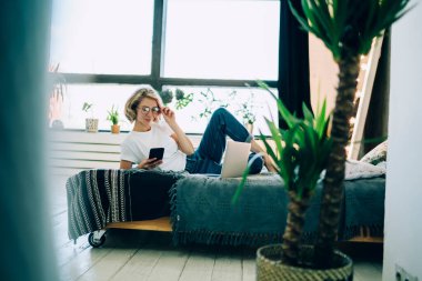 Gündüz vakti yatak odasında açık dizüstü bilgisayarla yatarken gözlüklü, akıllı telefonlu, güler yüzlü genç bir kadın.