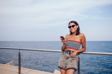 Gülümseyen milenyum kadınının yarım boy portresi. Elinde dijital hareket teknolojisi olan, yaz gezintisi sırasında kameraya bakan cep telefonu cihazlı mutlu Latin turist.