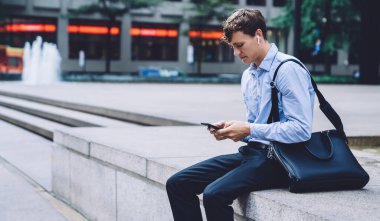 Resmi kıyafetleri ve kablosuz kulaklıkları olan bir erkek çalışan New York caddesinde çeşmenin kenarında telefonla mesajlaşıyor.