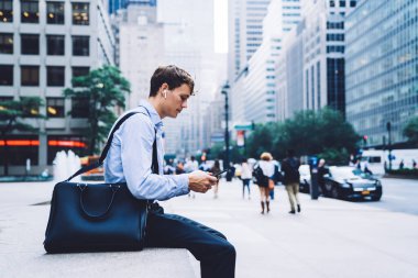 Klasik mavi tişörtlü ve kablosuz kulaklıklı bir erkek çalışanın New York 'taki yüksek binaların arka planında telefonla mesajlaşması.