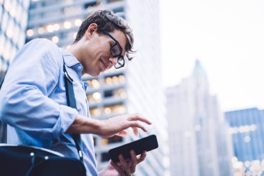 Gömlekli, kollu, gözlüklü, çantalı genç bir ofis çalışanı New York 'un arka planında telefonla gülümsüyor.