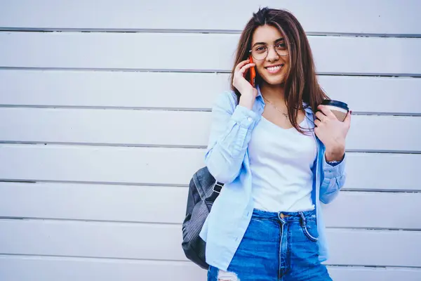 Portrait of cheerful brunette caucasian woman talking on mobile phone standing near wall with coffee to go, smiling hipster girl in stylish outfit enjoying smartphone conversation in roaming