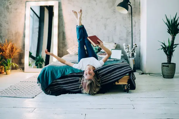 Comfy loft design interior in flat apartment, portrait of successful female reader holding bestseller book in hand and laughing feeling happiness from own life, cheerful woman lying on bed in home
