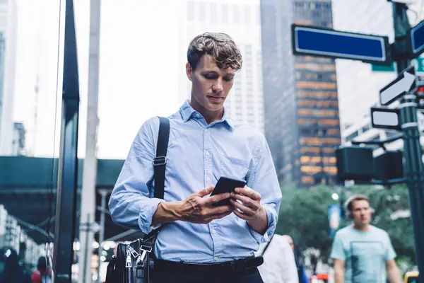 Intelligent businessman in classic outfit holding leather bag standing browsing on smartphone front of commercial buildings on street of New York city looking away
