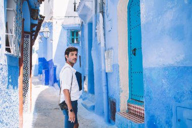 Fas 'ın mavi turistik şehrinde turistik gezintiler sırasında kameraya bakarken günlük giysiler içinde odaklanmış genç bir gezginin yan görüntüsü.