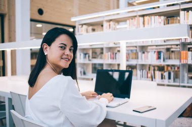 Başarılı kadın yöneticinin portresi kütüphanede oturmuş dizüstü bilgisayarla iş başında yazarken gülümsüyor başarılı bir iş kadını kameraya bakıyor ve modern teknolojileri araştırma ve iş için kullanıyor.