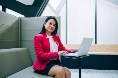 Kırmızı ceketli, gülümseyen beyaz bir kadın modern ofiste bilgisayarında yazı yazıyor. İnternette araştırma yapıyor. Olumlu bir kadın yönetici kameraya bakıp iletişim için teknoloji ve kablosuz internet kullanıyor.