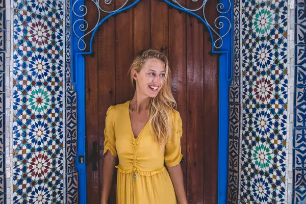 Glad smiling young gorgeous woman in casual clothes traveling in city exploring Morocco standing in old colorful street looking away